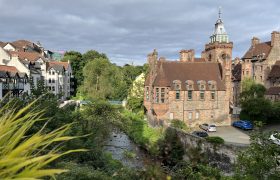Man sieht einen Fluss, der mit Bäumen und grünen Sträuchern gesäumt ist. Am rechten Ufer führt eine Straße entlang, neben der ein großes, zweistöckiges, braunes Gebäude steht, es hat eine dunkelbraunes Satteldach und einen Glockenturm. Es mutet wie eine Kirche an. Neben der Kirche und auf der anderen Flussseite stehen mehr alt aussehende Gebäude, die vornehmlich weiß sind. Sie haben allerdings dunkelrote Dächer. Vom Gesamtbild sieht es wie eine mittelalterliche Kleinstadt aus.