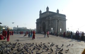 Gateway of India. Ein hohes Gebäude am Hafen von Mumbai steht auf der rechten Seite des Bildes. Davor stehen einige in rot gekleidete Mönche und es sind viele Tauben im Vordergrund zu sehen.