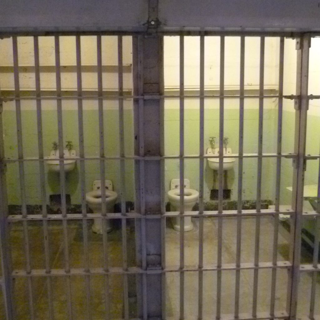 Eine Gefängniszelle mit Waschtisch, Sitzmöglichkeit und Toilette. Sehr karg.