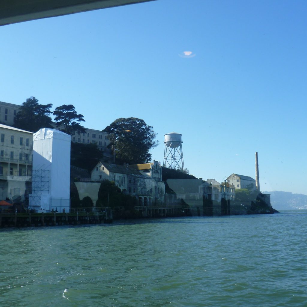 Die Insel vom Wasser aus. Man sieht neben einem Wasserturm auch Bäume, Gefängnismauern und Wachtürme.