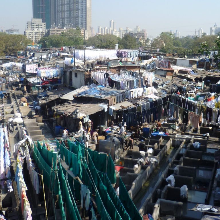 Dhobi Ghat. Wahrscheinlich die größte Outdoor-Wäscherei der Welt. Sie liegt in Mumbai. Man sieht viele Wäscheleinen, einige Parzellen, die mit Wasser gefüllt sind und in denen Arbeiter Wäsche waschen. Es sind auch einige Hütten mit Stoffdach zu erkennen.