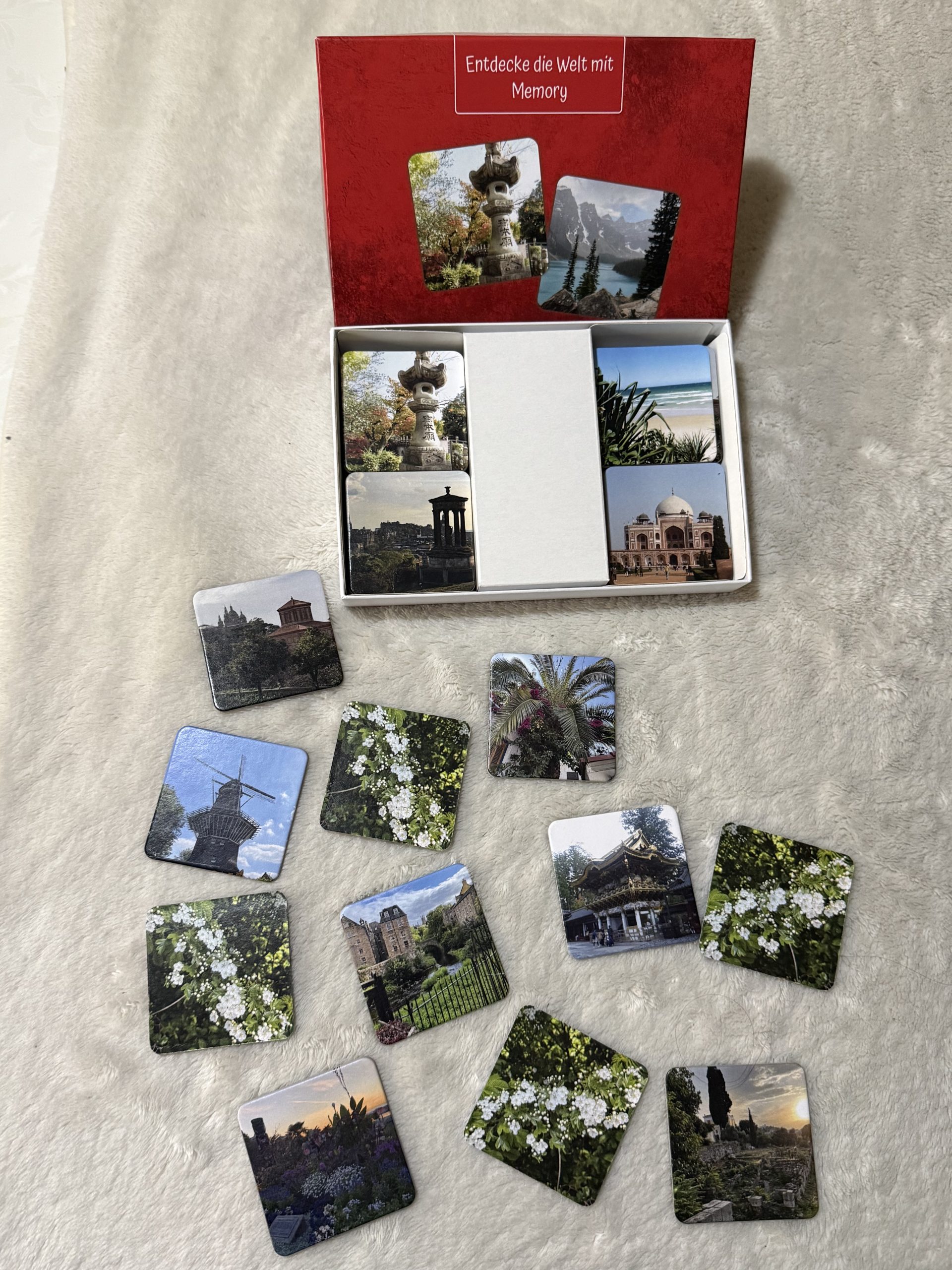Ein Memory Spiel mit Bildern aus Destinationen weltweit. Die Schachtel ist rot und man sieht viele kleinere Karten davor auf einer hellen Oberfläche verteilt liegen.