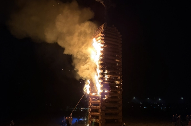 A wooden tower engulfed in flames. It stands in a meadow, and it is night.