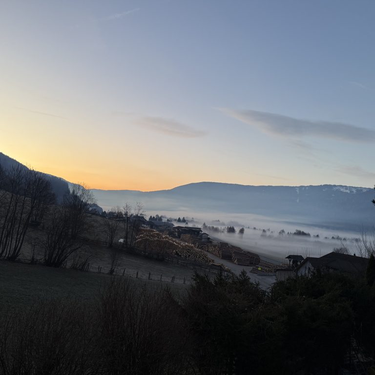 Eine Morgenstimmung in einer Berglandschaft. Man sieht die aufgehende Sonne, viele Berge und etwas Nebel im Tal liegen. Nur wenige Schleierwolken sind zu sehen.