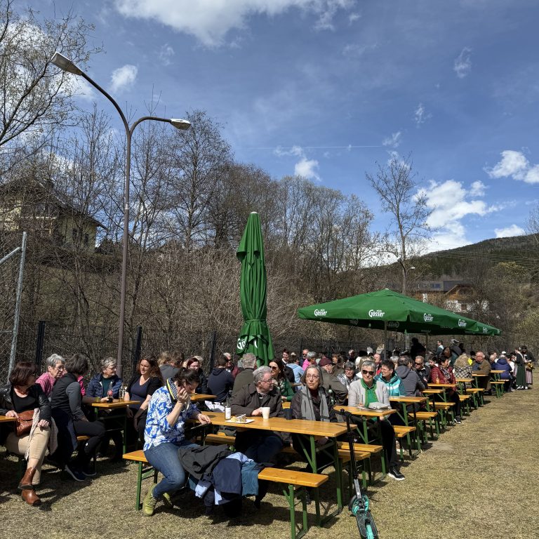 Man sieht einige Bierbänke mit Zuschauern unter blauem Himmel. Auch einige Bäume sind im Hintergrund zu sehen. Ein großer grüner Sonnenschirm ist aufgespannt.