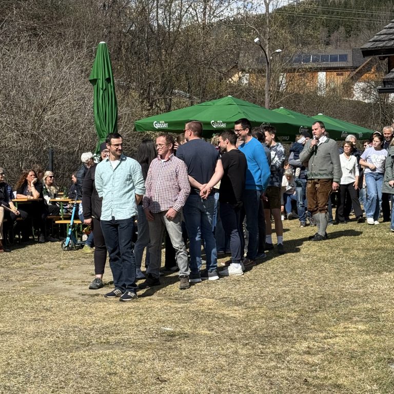 Man sieht eine Menge Menschen hintereinander auf einer Wiese stehen. Sie ist eher vertrocknet. Im Hintergrund sind Bäume zu sehen, einige grüne Sonnenschirme und Zuschauer.