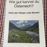 Ein Buch mit dem Titel "Wie gut kennst du Österreich". Auf der Vorderseite sieht man eine Alm mit einer Kuh. Das Ringbuch liegt auf einer rot weiß karierten Oberfläche.