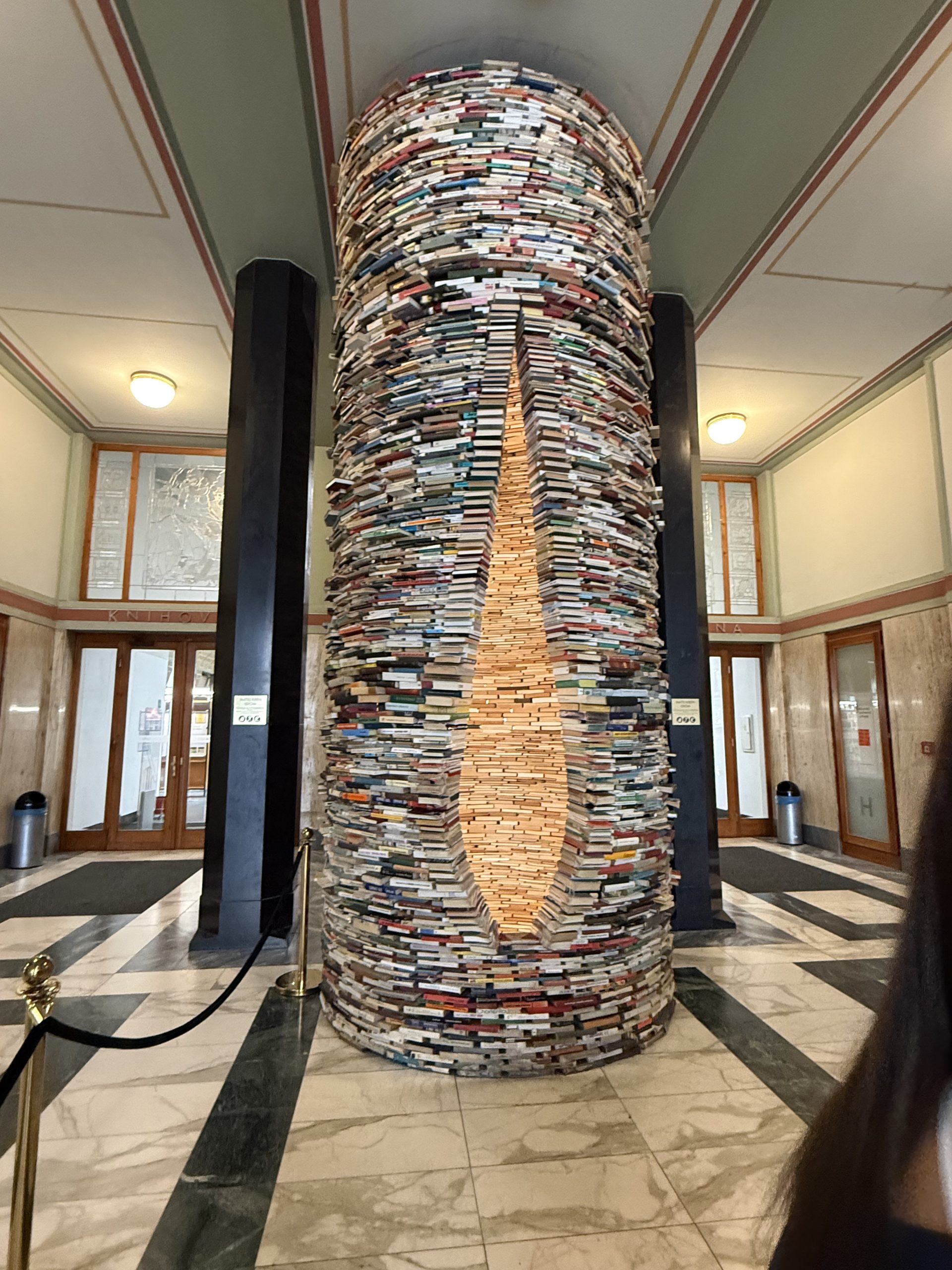 Ein Turm mit einer längstöffnung durch die man hineinschauen kann. Der Turm besteht aus geschichteten Büchern und steht in einem Raum. Er ist hell und freundlich. Der Boden ist gefliest und an den Wänden sind Holzverkleidungen angebracht.