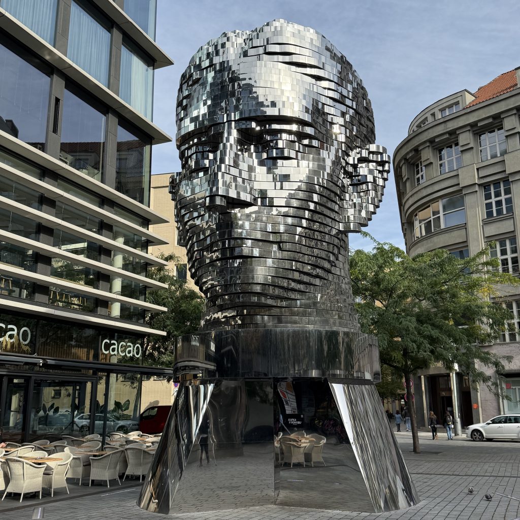 Man sieht einen riesigen, silbernen Kopf der aus mehreren Platten besteht. Er steht auf einem glänzenden Sockel. Links davon steht ein modernes Haus, auf der rechten Seite ein älteres Gebäude.