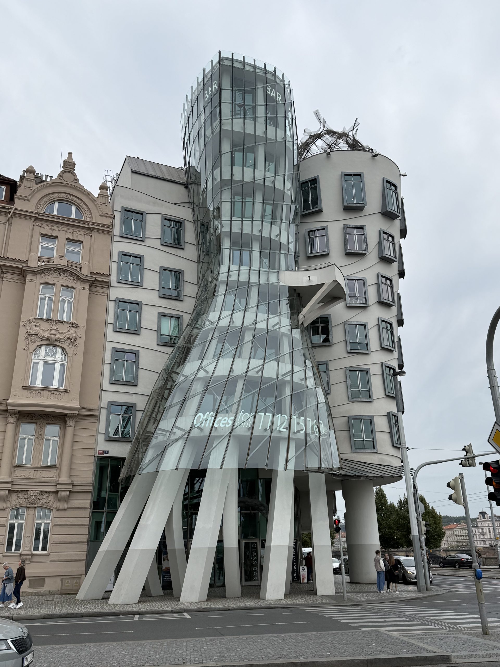 Ein mehrstöckiges Haus auf mehreren Stelzen. Es scheint zu wanken. Mittig ist es schmal und wird nach oben und unten etwas breiter. Es steht mittig zwischen zwei weiteren Gebäudeteilen und scheint sich im Wind zu bewegen. Die Front ist aus Glas und es ist Ewas in sich gedreht. 