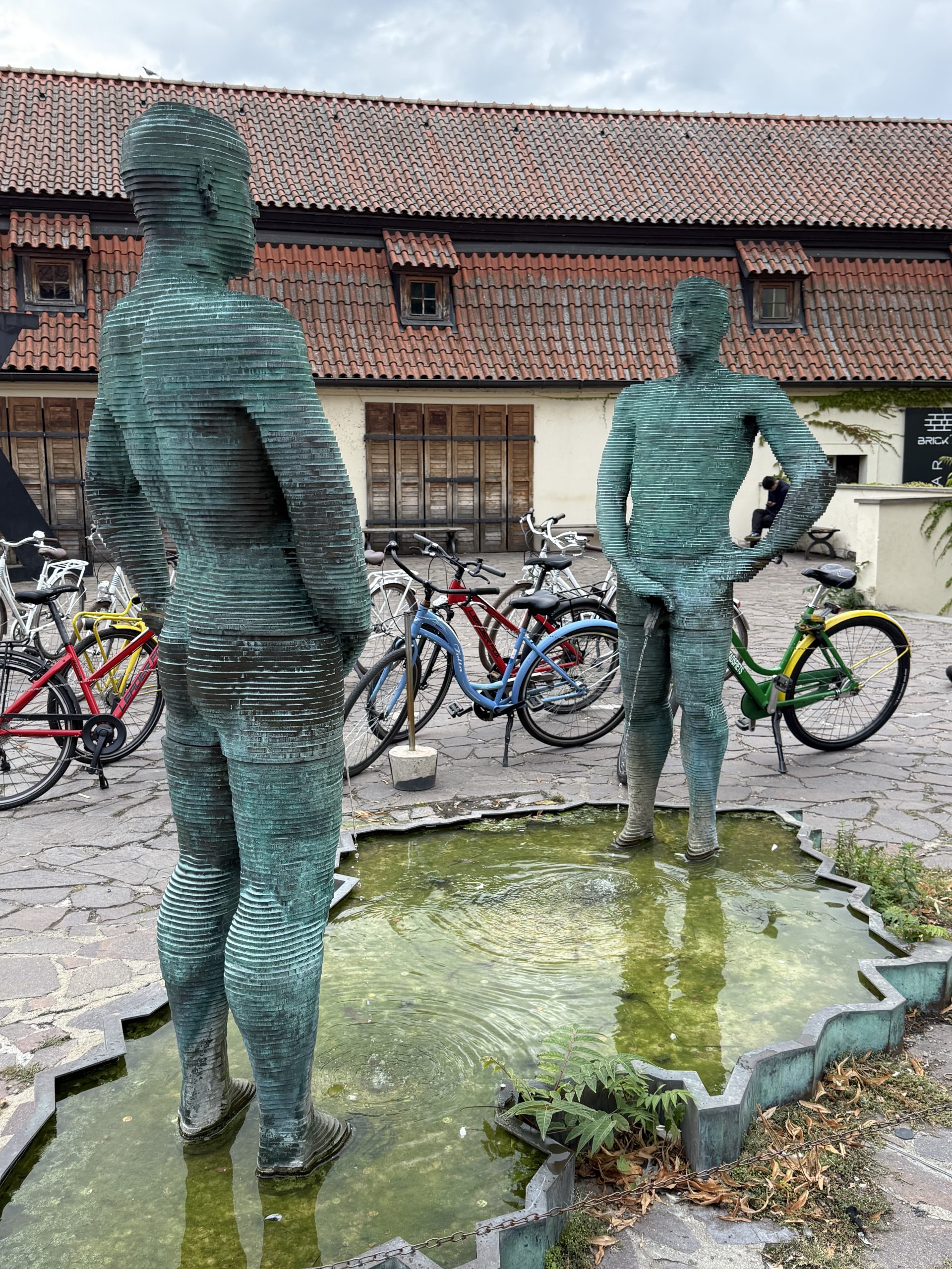 Bei diesem Kunstwerk sieht man zwei unbekleidete Männer aus grün angelaufenen Metall, die in einem Brunnen stehen. Sie urinieren in den Brunnen. Im Hintergrund sieht man Fahrräder von Besuchern und ein Gebäude