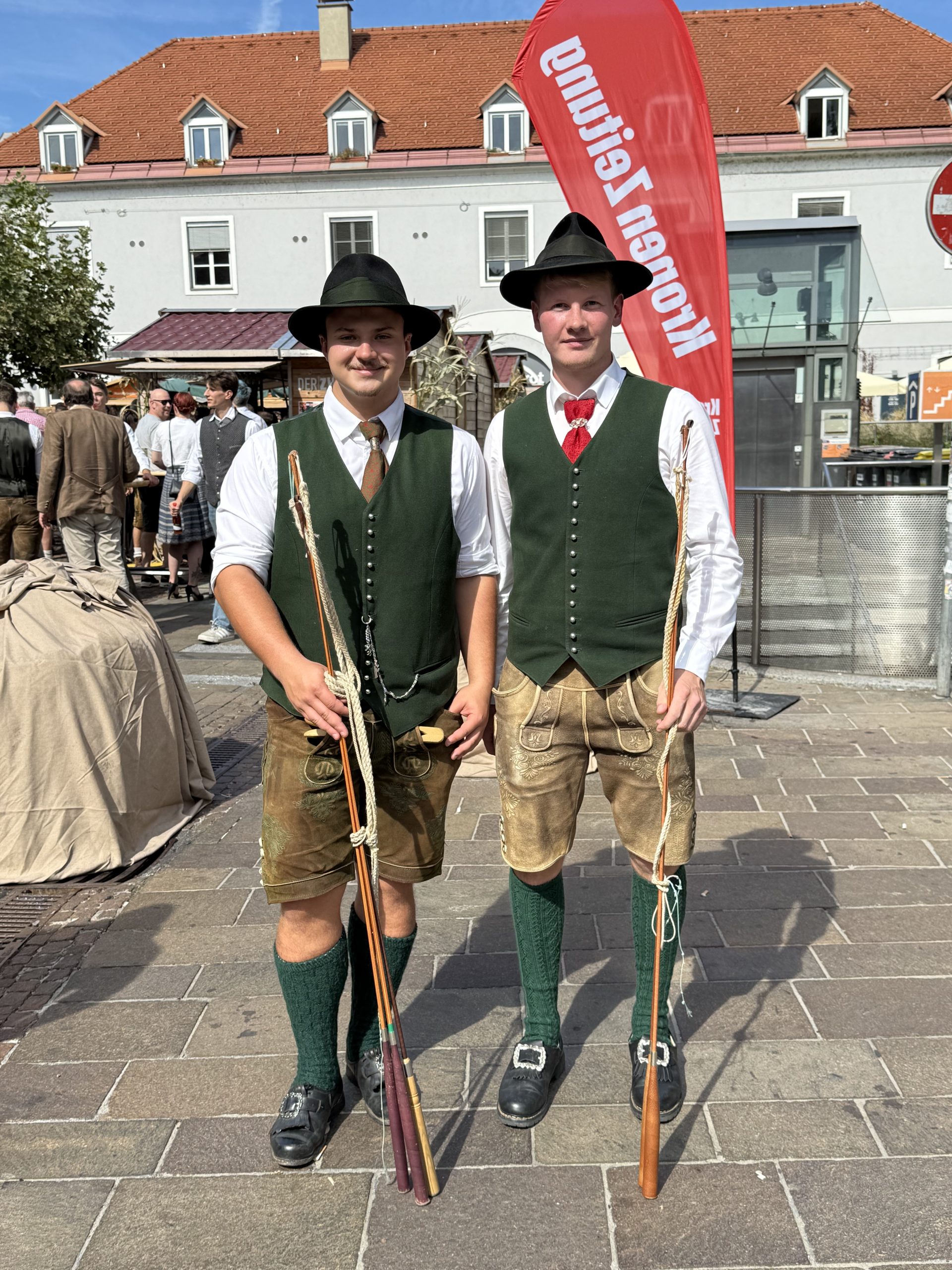 Zwei Männer in Tracht haben einen langen Stab in Händen. Sie tragen Hüte mit Gamsbärten, grüne Jacken über weißen Hemden und je eine rote Krawatte. Sie haben Lederhosen an und grüne Strümpfe. Sie stehen an einem großen Platz und lachen in die Kamera.