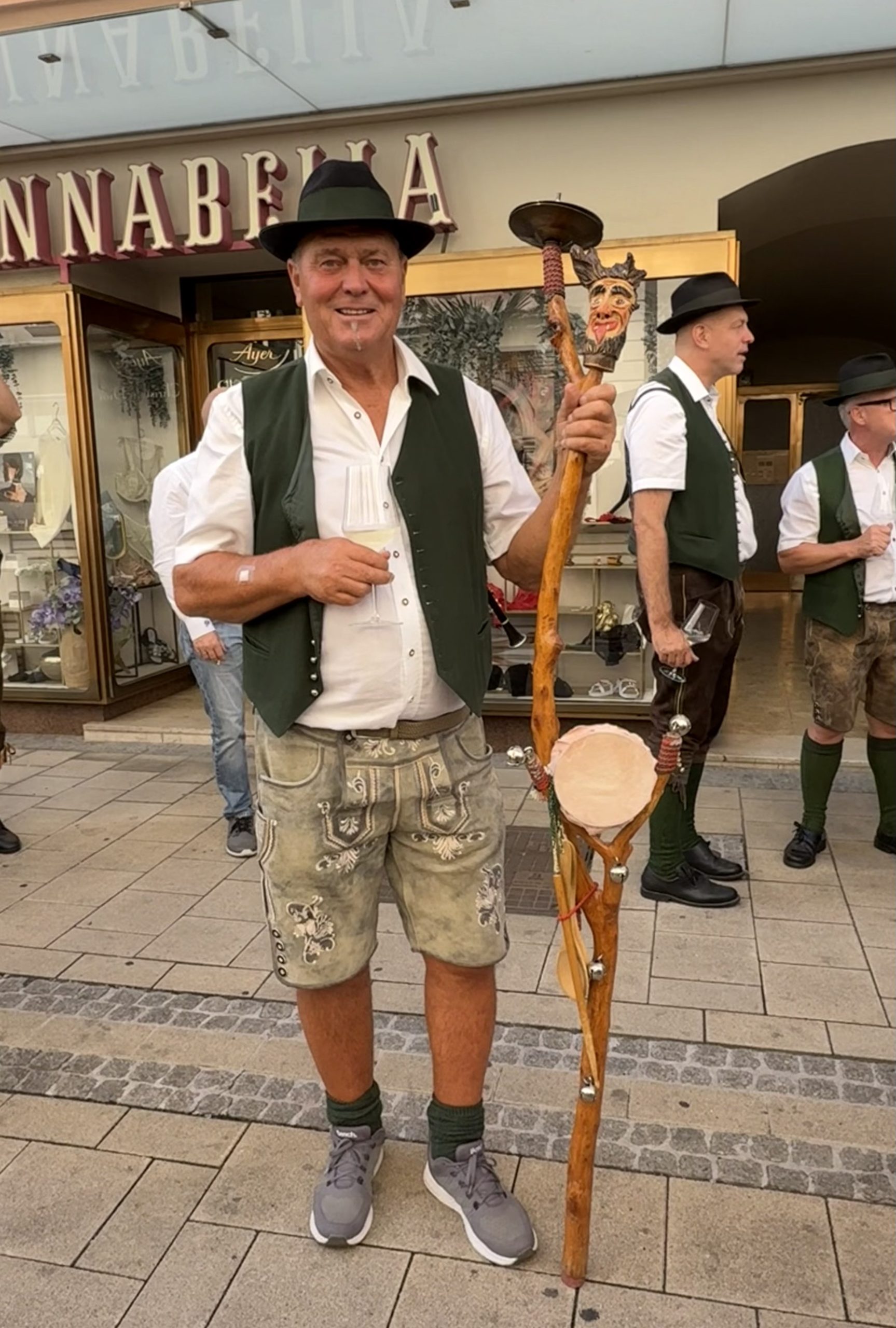 Ein Teufelsgeiger posiert für ein Foto. Er hat eine kurze Lederhose an, weißes Hemd und grüne Weste. Er trägt einen Hut, sieht direkt in die Kamera und hat sein Instrument in der rechten Hand. Er steht auf der Straße
