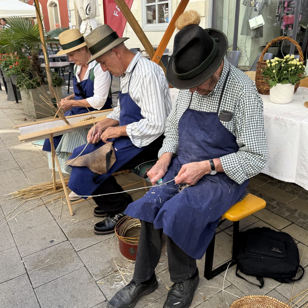 Drei ältere Herrschaften die mit blauen Schürzen auf Stühlen sitzen. sie sehen nach unten uns sind konzentriert. In den Händen haben sie lange dünne Halme. Alle drei tragen verschiedenfarbige Hüte.