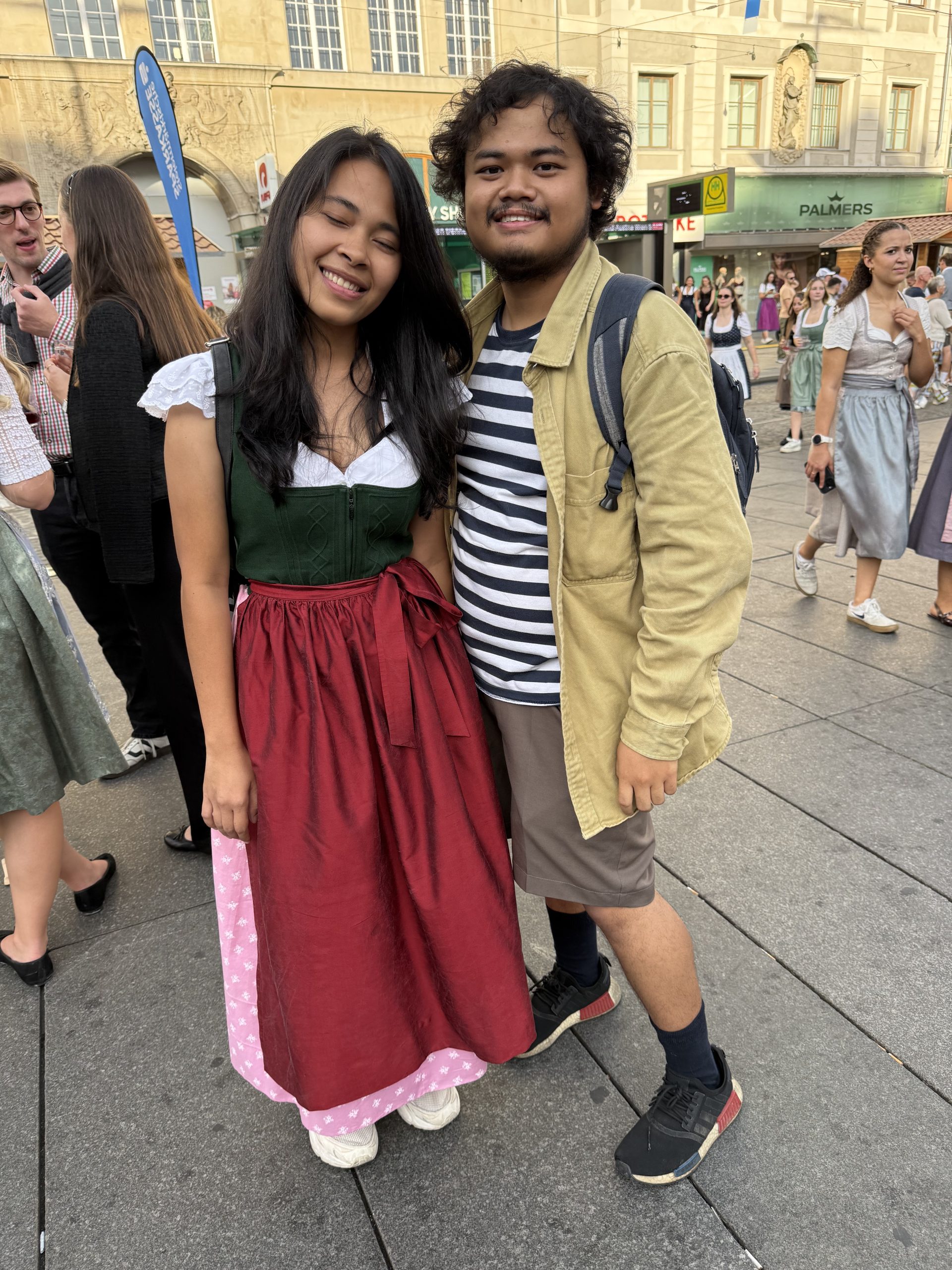 Rechts eine Dame im Dirndlkleid mit dunklen, langen Haaren, einer weißen Dirndlbluse und grünem Dirndl mit Roter Schürze. Die Bluse ist kurzärmelig. Daneben seht ein Mann mit leichtem Oberlippenbart und dunklen Haaren. Er hat eine beige Jacke an, darunter ein auergestreiftes Shirt und eine braune Hose. Er trägt einen Rucksack. Beide stehen auf einem belebten Platz und lachen in die Kamera.