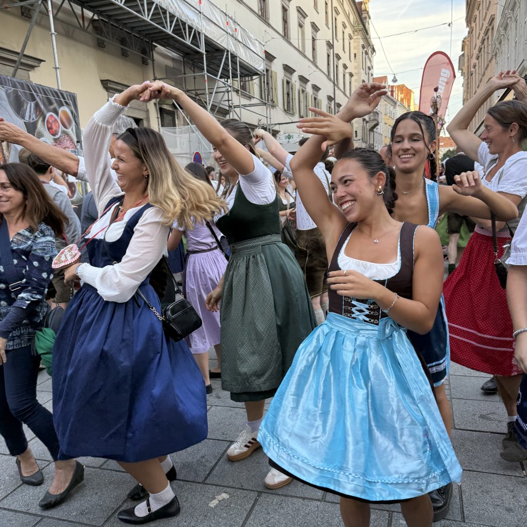 Junge Mädchen tanzen und drehen sich gegenseitig im Kreis. Sie tragen dabei Tracht. Sie tanzen auf den Straßen einer großen Stadt in einer großen Gruppe