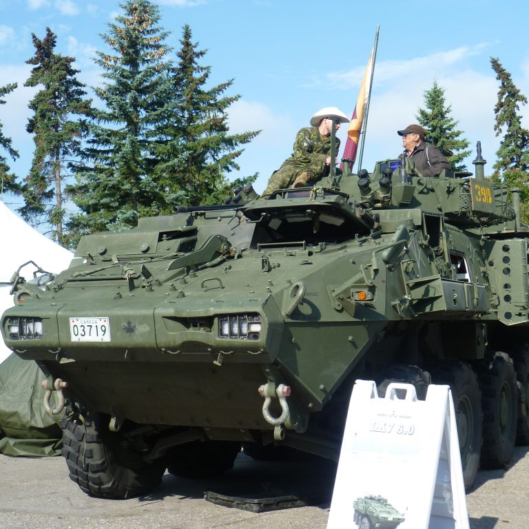 Ein grüner Panzer mit zwei Menschen die obenauf sitzen. Im Hintergrund sieht man Bäume. Der Panzer steht auf Asphalt.