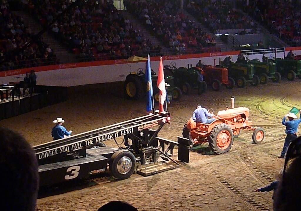 Traktor Pulling. Hier sieht man einen orangefarbenen Traktor der hinten ein langes Gestell angehängt hat. Es ist eine lange Rampe die auf Reifen montiert ist. Der Traktor fährt auf einem sandigen Untergrund. Im Hintergrund sieht man Zuschauer.
