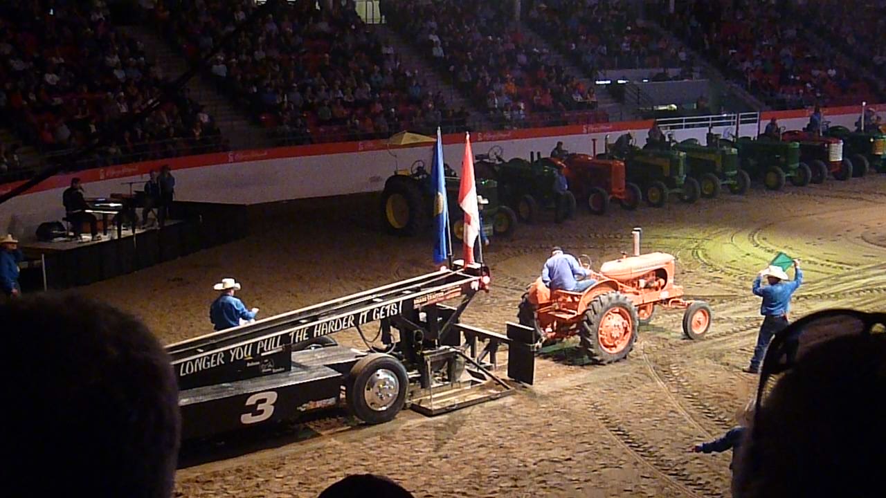 Traktor Pulling. Hier sieht man einen orangefarbenen Traktor der hinten ein langes Gestell angehängt hat. Es ist eine lange Rampe die auf Reifen montiert ist. Der Traktor fährt auf einem sandigen Untergrund. Im Hintergrund sieht man Zuschauer.