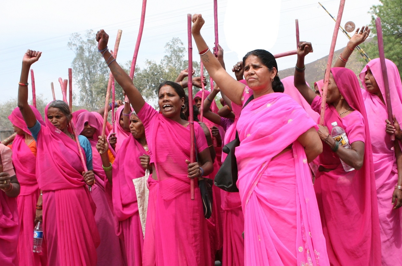 Man sieht Frauen in rosa Saris die eine Hand in die Luft strecken. Sie haben einen rot bemalten Stock in Händen und scheinen sehr entschlossen zu sein.