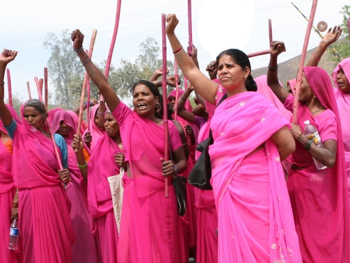 Man sieht Frauen in rosa Saris die eine Hand in die Luft strecken. Sie haben einen rot bemalten Stock in Händen und scheinen sehr entschlossen zu sein.