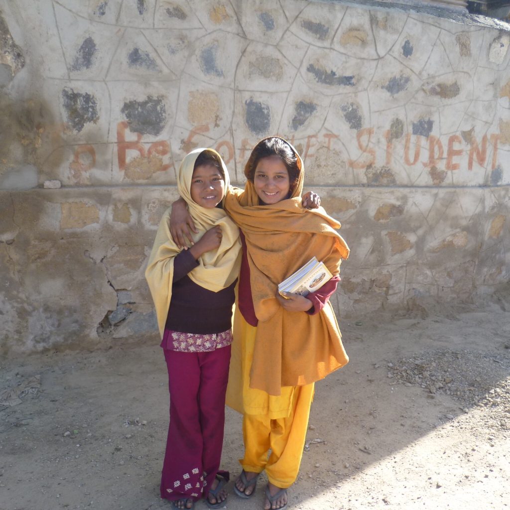 Zwei Mädchen posieren für die Kamera. Sie haben indische Kleidung an. Das Mädchen rechts trägt senffarbene Kleidung, das Mädchen links eine rote Hose mit verziertem Bund, darüber eine schwarze Kurta und einen goldfarbenen Schal, der teilweise ihre Haare bedeckt. Beide haben schwarze Haare, die nach hinten gekämmt sind. Sie stehen vor einer Steinmauer auf der ein Graffiti mit dem Wort "Student" steht.