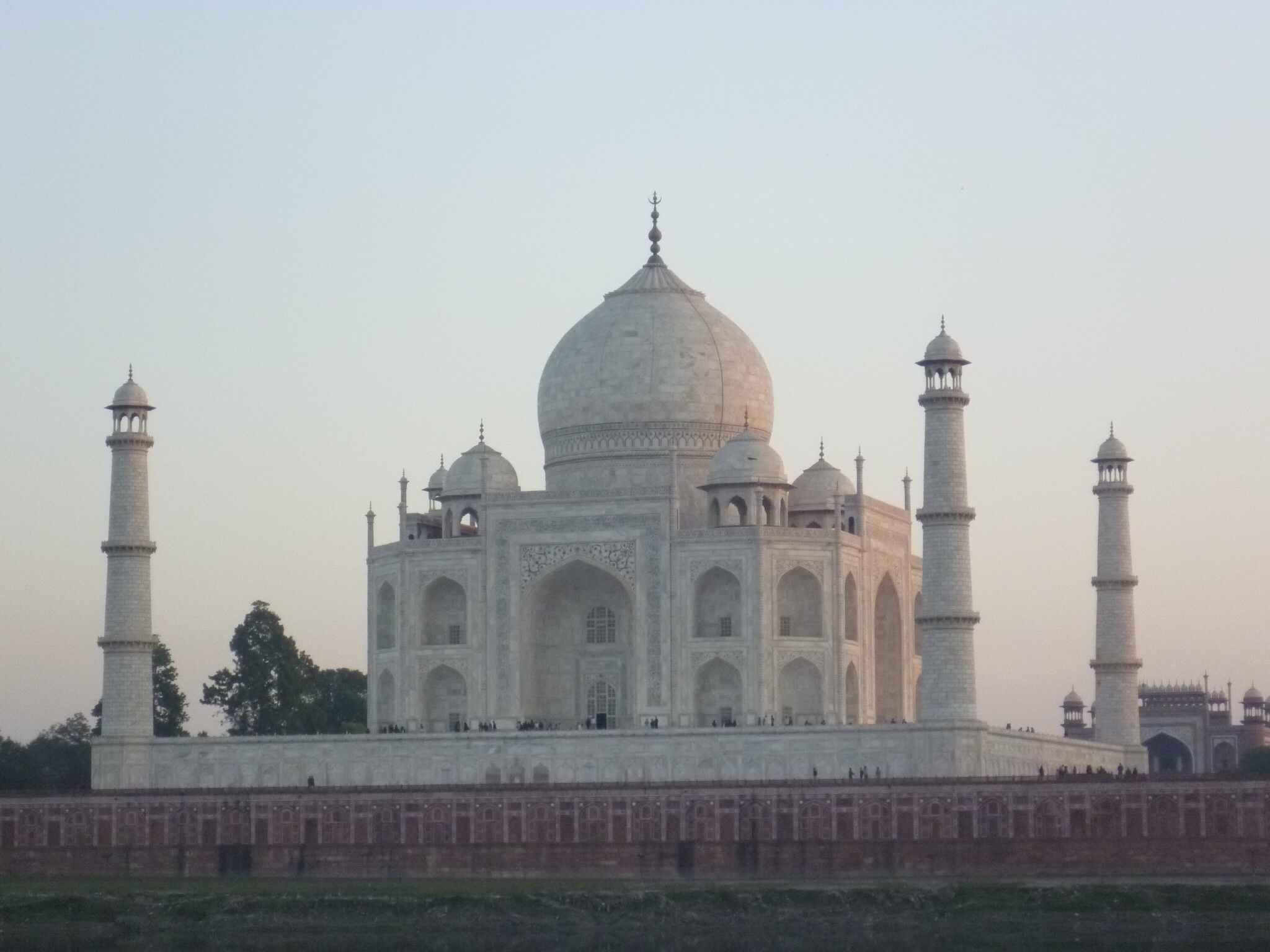 Das Taj Mahal bei Sonnenuntergang. Das Grabmal ist in rosafarbenes Licht getaucht. Man sieht es von der Ferne.